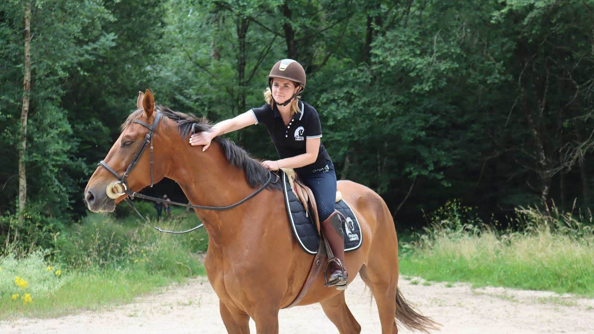 Une cavalière à l'arrêt carresse son cheval dans la forêt en souriant.